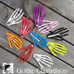 May include: Nine skeleton hand hair clips in various colors, including white, pink, purple, black, orange, red, blue, and yellow. The clips are arranged in a circle on a wooden surface.