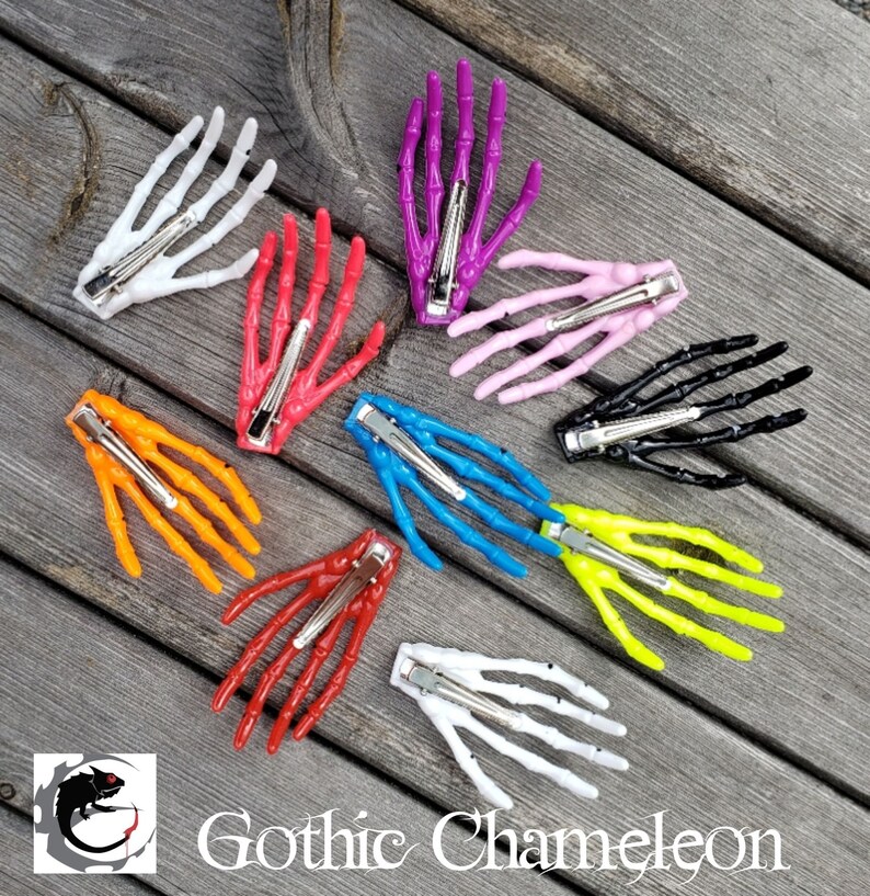 May include: Nine skeleton hand hair clips in various colors, including white, pink, purple, black, orange, red, blue, and yellow. The clips are arranged in a circle on a wooden surface.