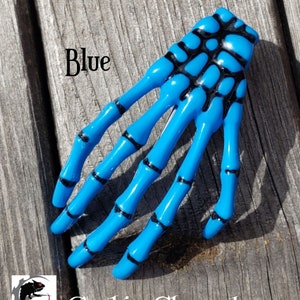 May include: Blue skeleton hand hair clip with black detailing.  The clip is made of plastic and has a shiny finish.