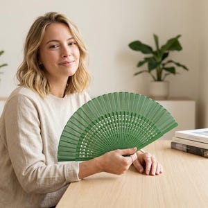 May include: A green folding hand fan. The fan is made of wooden slats and is held open. The fan is a solid green color. The fan is being held by a person sitting at a wooden table.