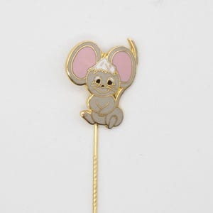 May include: A gold-toned stick pin featuring a cartoon mouse with gray fur, pink ears, and a white hat. The mouse is sitting upright. The pin has a gold-colored shaft and a cylindrical end.