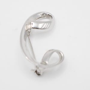 May include: A polished silver-toned metal ear cuff featuring a continuous, fluid, and twisted ribbon-like design that curves into an open loop shape. The surface is smooth and reflective, catching the light to highlight its curved, sculptural form.