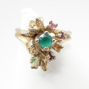 May include: A gold-toned ring with a central emerald-green gemstone and smaller surrounding gemstones in shades of pink, yellow, and green. The ring has a textured, organic design, resembling a cluster of leaves or branches.