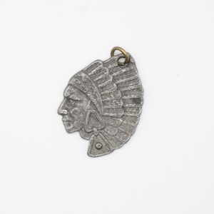 May include: A silver-colored pendant featuring a profile of a Native American chief wearing a headdress. The pendant has a textured surface and a small brass-colored loop at the top for hanging. The design is detailed and the pendant is set against a white background.
