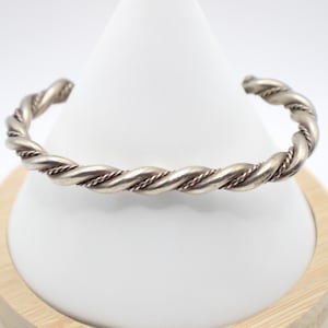 May include: A silver-toned cuff bracelet with a twisted rope design. The bracelet is displayed on a white cone-shaped stand with a wooden base, set against a white background. The cuff is a simple, yet elegant piece of jewelry.