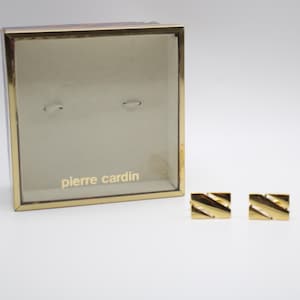 May include: A pair of gold-toned rectangular cufflinks with a diagonal grooved pattern, presented alongside a square display box featuring a gold-colored frame and the text pierre cardin printed in gold on the interior base of the box.