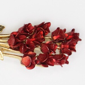 May include: A gold-toned metal brooch featuring a bouquet of red roses. The roses are clustered together, with detailed petals. The brooch has a curved, textured base with gold leaves.