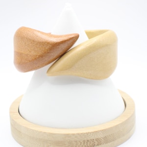 May include: Two wooden rings, one light brown and one medium brown, are displayed on a white cone-shaped stand with a bamboo base. The rings are interlocked, showcasing a natural wood grain.