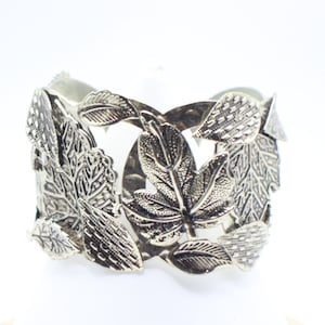 May include: Silver-toned cuff bracelet featuring a detailed leaf design. The leaves are intricately sculpted, creating a textured and naturalistic appearance. The bracelet has a wide band and a secure clasp, suitable for various wrist sizes.