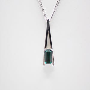May include: A silver-toned pendant necklace featuring a slender, triangular frame with a polished finish. At the base of the frame, a single rectangular, emerald-green gemstone is securely held by four prongs. The pendant hangs from a delicate silver chain.