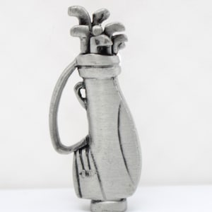 May include: A silver-colored golf bag pin with a handle and a set of golf clubs. The bag has a textured surface and is standing upright. The pin is a miniature representation of a golf bag.