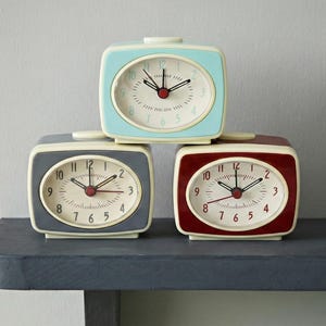 Pode incluir: Três despertadores retrô empilhados em uma prateleira. Os relógios são quadrados com cantos arredondados e apresentam um esquema de cores azul claro, cinza e bordô. Cada relógio tem um mostrador creme com algarismos pretos e ponteiros vermelhos.