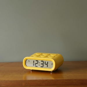 May include: A bright yellow digital alarm clock showing 12:34 PM. The clock has a rectangular display and a rounded, button-covered top. It is placed on a wooden surface.
