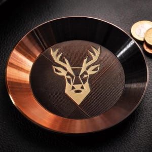 Könnte beinhalten: Ein runder Metalluntersetzer mit kupferfarbenem Rand und schwarzer Mitte. In der Mitte befindet sich ein goldfarbenes geometrisches Hirschkopfdesign. Rechts vom Untersetzer sind mehrere Münzen zu sehen, die auf einer schwarzen Lederoberfläche liegen.