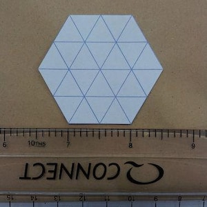 May include: A white paper hexagon with blue triangle grid lines, placed on a brown surface. A clear ruler with black markings and the word "CONNECT" is below the hexagon. The ruler measures in centimetres.