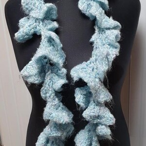 Puede incluir: Una bufanda de ganchillo de color azul claro con bordes con volantes. La bufanda se muestra en un maniquí negro.