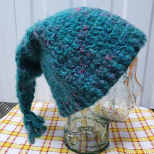 以下が含まれることがあります： 長い先のとがった尻尾が付いたターコイズブルーのニット帽。帽子はふわふわした質感で、ピンクと紫の糸が混ざっています。