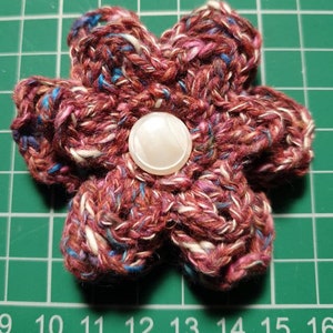 Puede incluir: Una flor de ganchillo con un botón blanco en el centro. La flor está hecha con una mezcla de hilo rojo, rosa, blanco y azul.