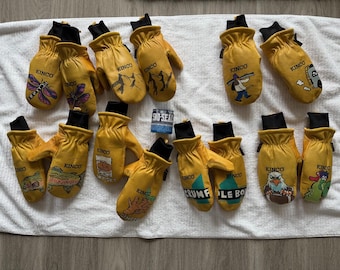 May include: A collection of yellow Kinco work mittens with black cuffs, featuring various embroidered designs such as a dragonfly, mountains, and a fisherman. A small can of Sko-Seal is also present.