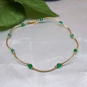 May include: A delicate gold bracelet with alternating emerald green and teal-colored beads. The bracelet is made of thin, curved gold bars and has a small clasp. The jewelry is displayed on a white, textured surface.