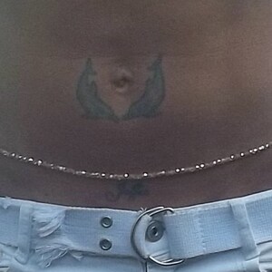 May include: A close-up of a woman's waist wearing a beaded waist chain with a white belt. The chain is made of small, clear beads and is worn over a white belt with silver buckle. The woman has a tattoo of two dolphins on her lower abdomen.