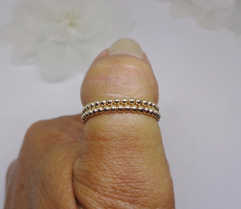 May include: Two gold beaded rings stacked on a finger. The rings are made of small, round beads.