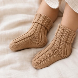 May include: A pair of tan-colored knit socks featuring a vertical ribbed pattern throughout the foot and ankle. The socks have a folded cuff at the top, providing a structured look. The knit texture is consistent, showing a clear stitch definition.
