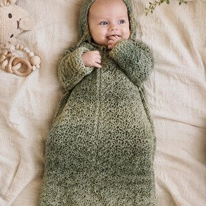May include: A sage green crocheted baby sleep sack featuring a hood and long sleeves. The garment is crafted with a textured star-like stitch pattern throughout, providing a consistent look from the hood down to the bottom hem of the sack.