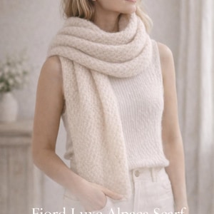 Op de afbeelding: Een vrouw draagt een crèmekleurige, getextureerde alpaca sjaal. De sjaal is om de nek en schouders gedrapeerd. De tekst "Fjord Luxe Alpaca Scarf" en "Variant I: Classic Luxe Scarf" zijn zichtbaar.