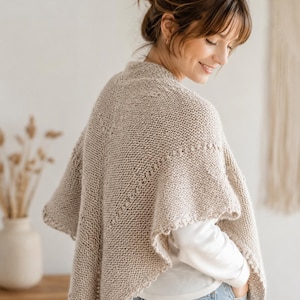 Könnte beinhalten: Ein beigefarbenes gestricktes Tuch mit einem strukturierten Kraus-rechts-Muster und einem dekorativen Bogenkantenabschluss. Das Kleidungsstück liegt über den Schultern und zeigt eine dreieckige Form mit dezenten vertikalen Details entlang der hinteren Mittelnaht.