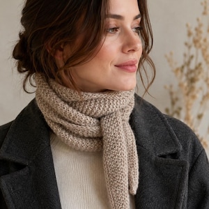 Könnte beinhalten: Ein beigefarbener Strickschal mit strukturiertem Strickmuster, der um den Hals gewickelt ist und über einem weißen Rippenpullover sowie einem dunkel anthrazitfarbenen Wollmantel getragen wird. Der Schal hat ein langes, spitz zulaufendes Ende.