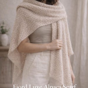May include: A light beige alpaca scarf, draped over the shoulders. The scarf is labelled "Fjord Luxe Alpaca Scarf" and "Variant III: Light Wide Wrap". The soft, textured wrap appears warm and comfortable, suitable for cooler weather.