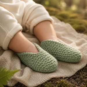 May include: A pair of sage green knit baby booties featuring a textured garter stitch pattern and a simple slip-on design with a V-shaped opening at the top. The booties are crafted from a soft yarn and cover the feet completely with a rounded toe.