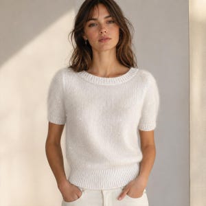 May include: A short-sleeved white knit jumper featuring a fuzzy, textured surface with subtle integrated sequins that catch the light. The garment has a round neckline and ribbed detailing along the collar, sleeve cuffs, and the bottom hemline.