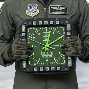 Fighter jet pilot wall clock in the style of a MFD (multi function display). Square black housing with light grey buttons and green lcd displaying time.