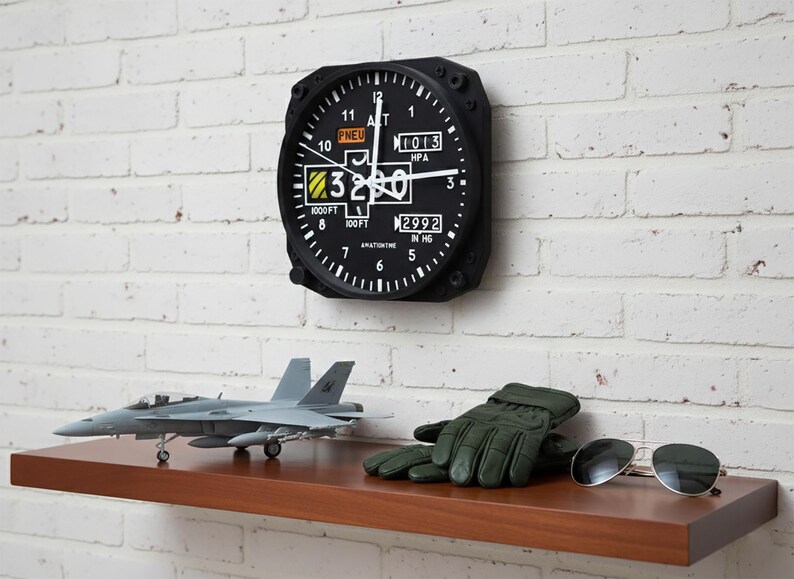 May include: Aviation-themed decor featuring a black altimeter clock on a white brick wall. Below, a model airplane, green gloves, and sunglasses rest on a wooden shelf. The clock displays numbers and text, including "1000FT" and "2992 IN HG".