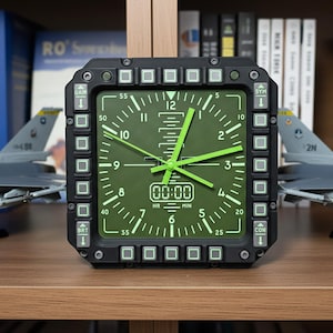 May include: A black and green analog clock with a square face, featuring green hands and numerals. The clock has a digital display showing "00:00". The clock is surrounded by buttons and is set against a backdrop of books and model airplanes.