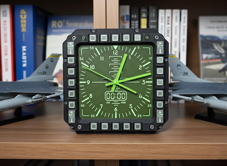 May include: A black and green analog clock with a square face, featuring green hands and numerals. The clock has a digital display showing "00:00". The clock is surrounded by buttons and is set against a backdrop of books and model airplanes.