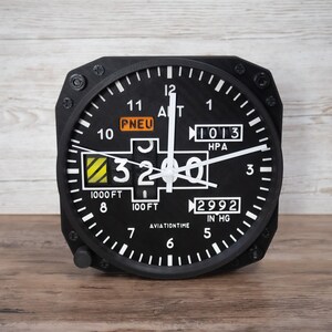 May include: Black aviation-themed clock with white numerals and hands. The clock face displays altitude and pressure readings, with the text "AVIATIONTIME" at the bottom. The clock is set to 3200 feet.