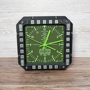 Fighter jet pilot wall clock in the style of a MFD (multi function display). Square black housing with light grey buttons and green lcd displaying time.