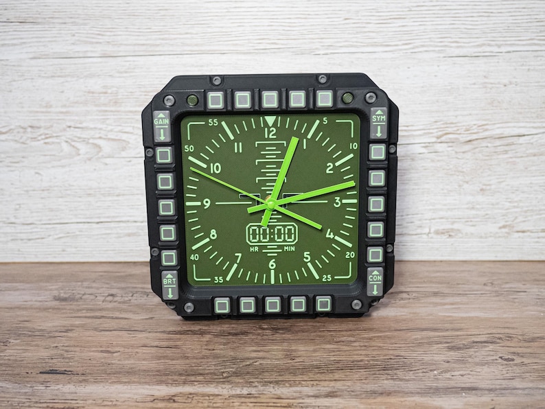 Fighter jet pilot wall clock in the style of a MFD (multi function display). Square black housing with light grey buttons and green lcd displaying time.