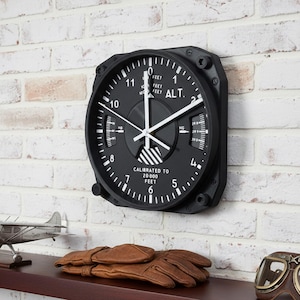 May include: Black aviation-style wall clock with white details, fixed to a white brick wall. The clock face displays "ALT" and "CALIBRATED TO 20,000 FEET". A model aeroplane, leather gloves, and aviator goggles are on a shelf below.