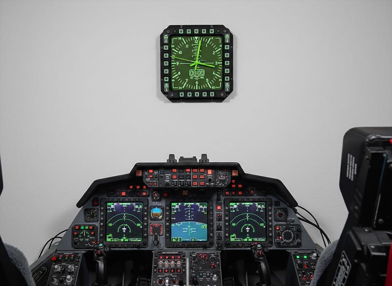 Fighter jet pilot wall clock in the style of a MFD (multi function display). Square black housing with light grey buttons and green lcd displaying time.