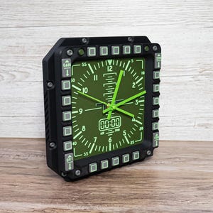 Fighter jet pilot wall clock in the style of a MFD (multi function display). Square black housing with light grey buttons and green lcd displaying time.