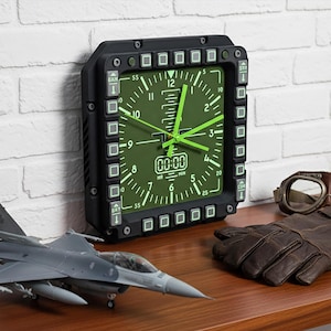 May include: Aviation-themed display with a black and green clock, model jet, brown leather gloves, and vintage aviator goggles. The clock has a square face with green hands and a black frame. The items are on a wooden surface.