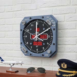 May include: Aviation-style wall clock with a grey frame and black face, featuring white hands and red digital displays. The clock is mounted on a white brick wall, with a model aeroplane, sunglasses, and a pilot's hat on a wooden surface below.