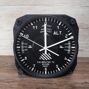 Pilot gift wall clock modeled after a Cessna aircraft cockpit altimeter. This aviation decor features realistic flight instrument details, a black bezel, and white dial lettering and clock hands.