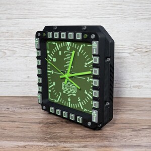 Fighter jet pilot wall clock in the style of a MFD (multi function display). Square black housing with light grey buttons and green lcd displaying time.