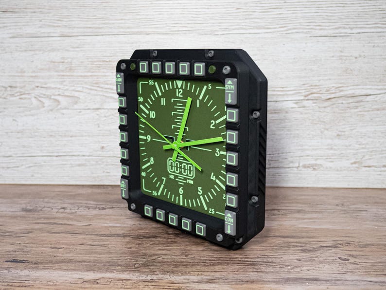 Fighter jet pilot wall clock in the style of a MFD (multi function display). Square black housing with light grey buttons and green lcd displaying time.