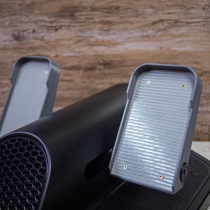 Può includere: Uno stepper nero e grigio con due pedali grigi. I pedali hanno una superficie a coste e sono attaccati a una base nera. Lo sfondo è una superficie di legno.